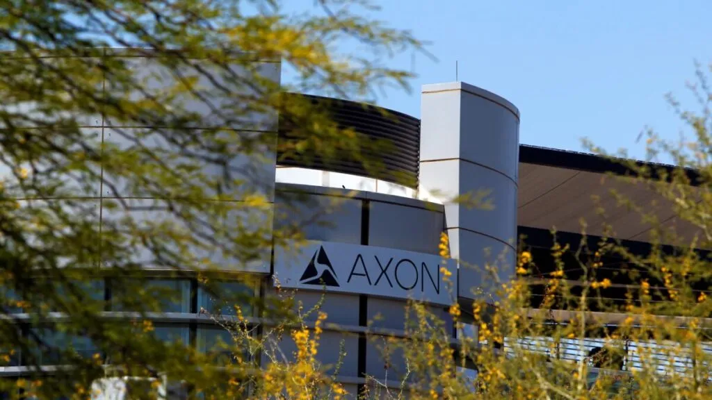 Fotografía editorial de una oficina moderna con pantallas mostrando software de seguridad y el logo de Axon en el fondo.