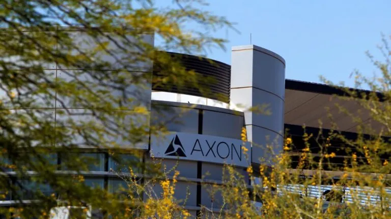 Fotografía editorial de una oficina moderna con pantallas mostrando software de seguridad y el logo de Axon en el fondo.