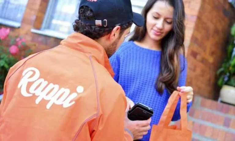 Fotografía editorial de un repartidor de Rappi en motocicleta con mochila naranja, recorriendo una ciudad latinoamericana moderna.