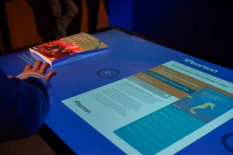 Persona interactuando con mesa digital táctil que muestra contenido educativo de ciencias de Pearson, integrando tecnología y aprendizaje aplicado en un entorno moderno