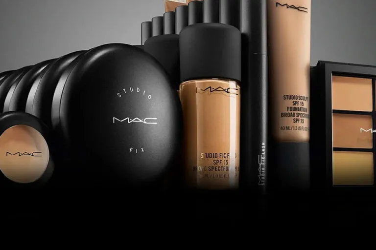 Productos icónicos de MAC Cosmetics en un mostrador de maquillaje con labiales, brochas y empaques negros característicos de la marca.
